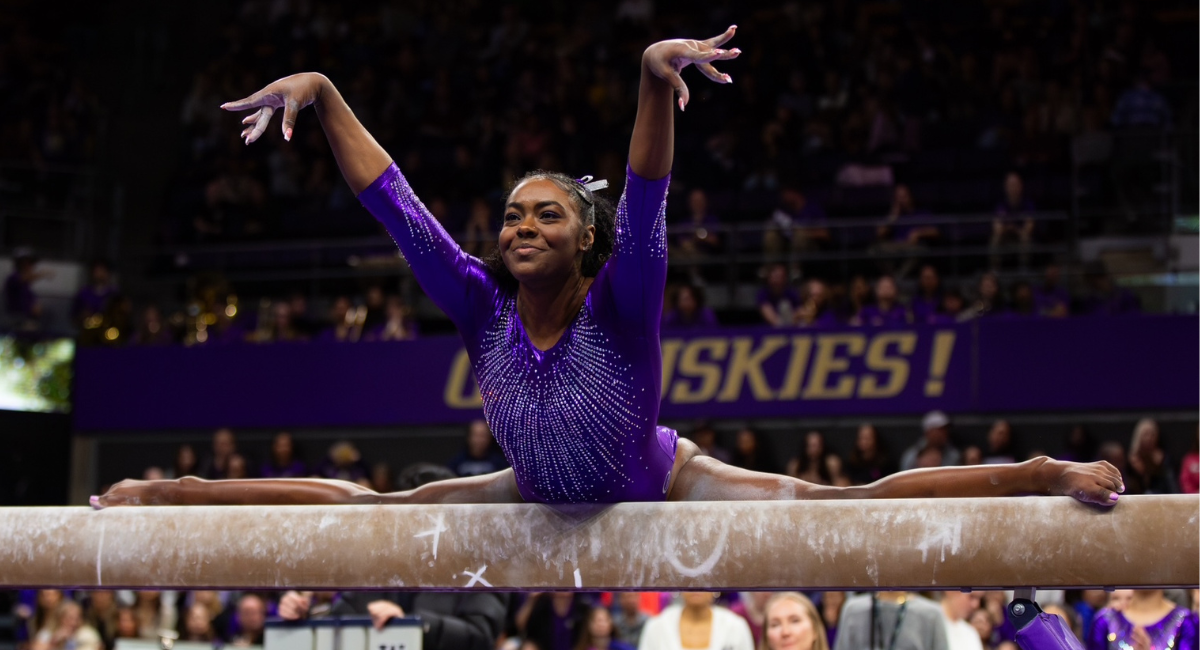 Deiah-Marie Moody performs the splits on a gymnastics beam and poses with her hands in the air.