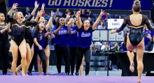 A group of UW gymnasts cheer on the sidelines as a gymnast walks across the mat toward them.